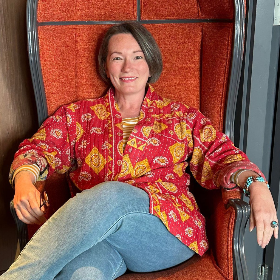 Middle-aged woman with brown hair sitting in orange chair and wearing a brightly colored jacket.